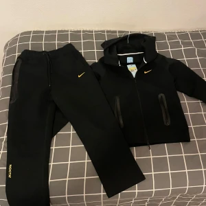 Svarta NOCTA mjukisbyxor från Nike - HELT NYA OANVÄNDA Svarta mjukisbyxor från Nike NOCTA med gul Swoosh och NOCTA-logga på benet. Byxorna har elastisk midja med snörning, svarta dragkedjor vid fickorna och detaljer i gult på baksidan. Sköna och stilrena, perfekta för chill eller träning.