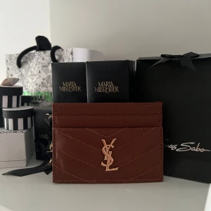 Brun YSL plånbok i skinn - Supersnygg brun plånbok från YSL i äkta skinn med guldigt YSL-logo framtill. Plånboken har flera kortfack och ett stilrent, quiltat mönster med snygga sömmar. Perfekt för dig som vill ha en lyxig och stilren accessoar i fickan.