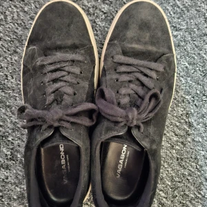Svarta mockasneakers från Vagabond storlek 40 - Svarta sneakers från Vagabond med ovandel i mjuk mocka och klassisk snörning. Skorna har en rund tå och vit platt sula med Vagabond-logga på hälen och undersidan. Perfekt för dig som gillar stilrena och enkla sneakers med en snygg kontrast mellan svart och vitt. Använda men i fint skick. Hör av dig vid minsta fundering! 😁