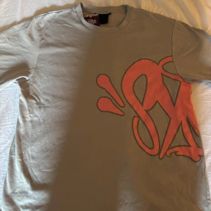 Grå t-shirt med röd graffiti från SYNA - Snygg grå t-shirt från SYNA med stort rött graffiti-tryck som sträcker sig över både fram- och baksidan. Klassisk rund hals och korta ärmar. Materialet är mjuk bomull. Skick är 10/10 aldrig använd 