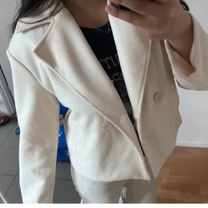 Beige kort kappa med knappar - Snygg beige kappa med klassisk krage och två knappar framtill. Jackan har en stilren och enkel design som passar perfekt till jeans eller kostymbyxor. Materialet känns mjukt och har en lätt struktur, vilket gör den bekväm att bära hela dagen.