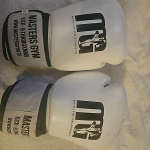 Vita boxningshandskar Masters Gym - Säljer ett par vita boxningshandskar från Masters Gym, perfekta för kick- och thaiboxning. Handskarna har svart logga och text på ovansidan samt breda muddar med kardborre för extra stöd. Snygg och stilren design som passar dig som vill träna hårt.