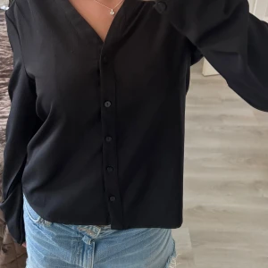 Svart v-ringad skjorta med knappar - Snygg svart skjorta med v-ringning och knappar hela vägen framtill. Långärmad modell med lite loose passform, perfekt till jeans eller kjol. Materialet känns mjukt och lätt, vilket gör den skön att bära. Enkel och stilren design som funkar till många olika looks. Använd fåtal gånger💕
