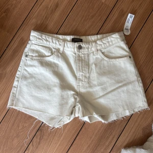 Beige jeansshorts från Lindex - Snygga beige jeansshorts från Lindex med rå kant och klassisk femficksdesign. Modellen är högmidjad och har bälteshällor samt knapp och dragkedja framtill. Materialet är en mjuk bomullsblandning som känns chill och passar perfekt till varma dagar.