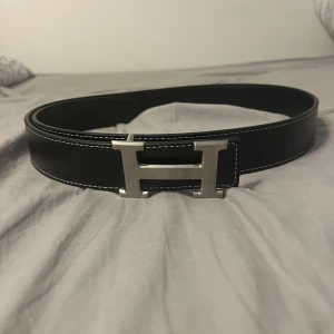 Svart H skärp - Snyggt svart skärp från H med ett stort H-format spänne i silverfärgad metall. Bältet är tillverkat i slätt skinn och har vit kontrastsöm längs kanterna. Perfekt accessoar för att lyfta din outfit med en lyxig touch.