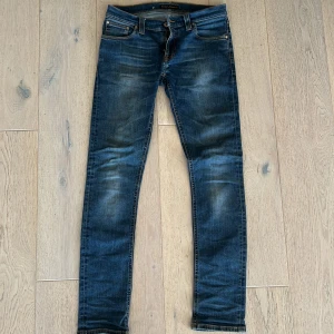 Blå slim fit jeans från Nudie Jeans (modell: tube Kelly) - Säljer ett par blå riktigt feta jeans från Nudie med klassisk femficksdesign och snygga slitningar. Jeansen har orangea sömmar och broderade bakfickor. Passar dig som gillar en smal siluett och vill ha ett stilrent och coolt plagg i garderoben, perfekt inför skolstarten. Priset går att diskutera vid snabb affär.