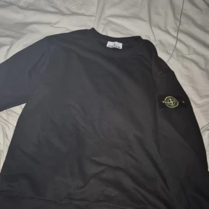 Svart sweatshirt från Stone Island  - Svart sweatshirt från Stone Island med klassisk rund hals och lång ärm i storlek Small. Ikonisk Stone Island patch sitter på vänster ärm. Tröjan är i mjuk bomull och har en clean, stilren look som passar perfekt till streetwear 
