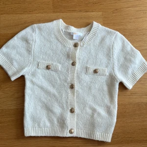 Vit kortärmad kofta från Gina Tricot - Supersöt vit kofta från Gina Tricot med korta ärmar och snygga guldfärgade knappar framtill. Koftan har två dekorativa fickor på bröstet och är stickad i mjukt material med ribbade kanter vid hals, ärmslut och nederkant. Perfekt för att ge din outfit en clean och fräsch vibe.   Den sticks inte utan den e bekväm och skön🤗