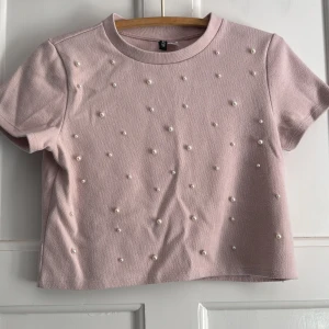 Ljusrosa croppad topp med pärlor H&M - Superfin ljusrosa croppad topp från H&M Divided, dekorerad med pärlor på framsidan för en extra söt vibe. Kortärmad och med rund hals, perfekt till jeans eller kjol. Mjukt material som känns skönt mot huden.