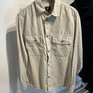 Beige overshirt från H&M med fickor - Snygg beige overshirt från H&M i storlek S. Skjortan har två stora bröstfickor med lock och knappar, klassisk krage och knäppning hela vägen fram. Tillverkad i mjuk bomull med långa ärmar och knappar vid manschetterna. Perfekt att styla lager på lager.