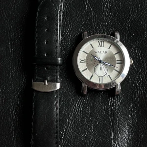 Klassisk silverfärgad Walar klocka - Stilren klocka från Walar med silverfärgat runt boett och svartvit urtavla med romerska siffror. Svart läderarmband med klassiskt spänne. Urtavlan har två små subdials och tydliga visare. Perfekt accessoar för dig som gillar tidlös design.