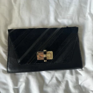 Svart kuvertväska från Diane von Furstenberg - Snygg svart kuvertväska från Diane von Furstenberg i skinn med diagonala ränder på framsidan och guldfärgat lås. Insidan har ett coolt mönster och en praktisk innerficka med dragkedja. Perfekt storlek för det viktigaste och stilren design