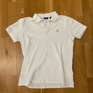 Vit pikétröja från GANT - Säljer en klassisk vit pikétröja från GANT i storlek S. Tröjan har en stilren design med korta ärmar och knappar framtill. Perfekt för både vardag och lite finare tillfällen. Passar bra till jeans eller chinos för en avslappnad men snygg look.