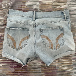 Ljusblå jeansshorts från Hollister - Säljer ett par ljusblå jeansshorts från Hollister med råa, fransiga kanter och klassiska fem fickor. Snygga detaljer med orange sömmar på bakfickorna och knapp samt dragkedja framtill. Perfekta för varma dagar och chill stil.