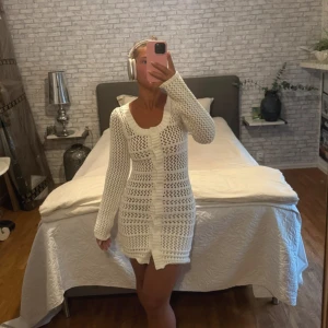 Vit stickad kortklänning med knappar - Supersöt vit stickad klänning med lång ärm och knappdetaljer framtill. Den är kort i modellen och har ett öppet, virkat mönster som ger en chill och trendig vibe. Perfekt för dig som gillar en feminin och fräsch stil.
