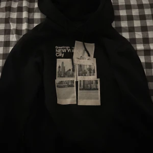 Svart hoodie med New York-print - Svart hoodie med coolt New York City-print på bröstet och fyra polaroidbilder av stadens skyline. Tröjan har huva med snörning och en avslappnad passform, perfekt för dig som gillar streetstyle och urban vibe.