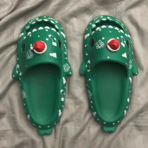 Gröna juliga krokodilsandaler - Unika gröna sandaler formade som krokodiler med vita julmotiv som granar, presenter och strumpor. Framtill sitter en röd tomteluva som extra detalj. Sandalerna har öppen tå och är tillverkade i syntetmaterial, perfekta för att sticka ut under julen. Det är lite sönder på tomteluvan som man ser på en av bilderna stl 44-45 men passar också 43