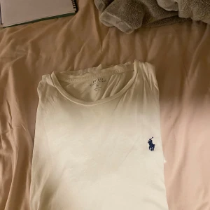 Vit t-shirt från Polo Ralph Lauren - Vit t-shirt från Polo Ralph Lauren med klassisk rund hals och blå broderad logga på bröstet. Modellen är kortärmad och har en clean, stilren look. Tillverkad i mjuk bomull som känns skön mot huden. Perfekt för dig som gillar enkel och snygg stil.