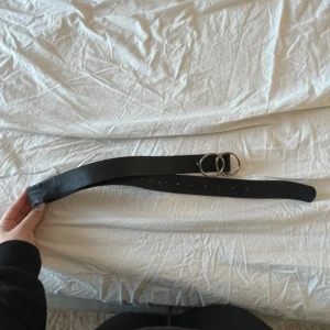 Svart bälte med dubbel ring-spänne - Snyggt svart bälte med stilrent dubbel ring-spänne i silverfärg. Bältet är brett och har flera hål för justering. Materialet är skinnliknande och passar perfekt till jeans eller kjol för en cool och modern look.