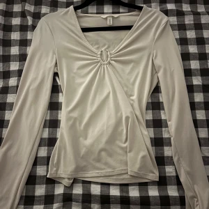 Ljusbeige långärmad topp från H&M - Snygg ljusbeige långärmad topp från H&M med v-ringning och cool rynkad detalj med ring framtill. Mjuk och stretchig polyester som sitter skönt och har en slim passform. Perfekt för dig som gillar stilrena och trendiga plagg.