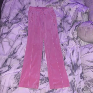 Rosa Juicy Couture mjukisbyxor med strass - Säljer ett par ikoniska rosa mjukisbyxor från Juicy Couture med glittrande strassdetaljer. Byxorna har bred resår i midjan, snörning och raka ben. På baksidan står det 'Juicy' i stora strassbokstäver och framtill finns JC-loggan i strass. Perfekt för dig som älskar Y2K-stilen.
