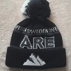 Svart och vit Åre mössa med tofs - Snygg svart och vit mössa med stor tofs på toppen och texten 'ÅRE' framtill. Upptill står det 'SWEDEN' och på uppviket finns ett broderat bergmotiv. Stickad design som passar perfekt för kalla dagar.