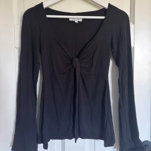 Svart långärmad topp från Design Si - Svart långärmad topp från Design Si i mjuk bomull. Toppen har en snygg v-ringning och cool knyt-detalj framtill som ger en trendig look. Passformen är normal och ärmarna är långa med lätt utsvängd form. Perfekt för dig som gillar stilrena plagg med en twist. Den är använd en del men är fortfarande i super skick.