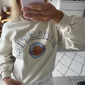 Beige sweatshirt Athletics från Gina Tricot - Säljer en beige sweatshirt från Gina Tricot med trycket 'ATHLETICS Madison Wisconsin United States' i blått och brunt framtill. Tröjan har rund hals, ribbade muddar och lång ärm. Perfekt för en chill vibe.