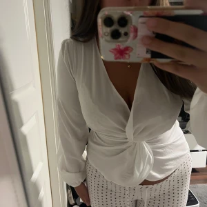 Vit omlottblus  - Säljer en fräsch vit omlottblus från NLY One i storlek S. Blusen har lång ärm, djup v-ringning och snygg knutdetalj framtill som ger en cool twist. Materialet är mjukt och lätt, perfekt för att styla med jeans eller kjol. Passformen är normal och blusen är enkel att matcha.