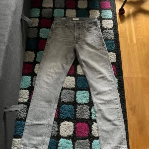 Grå jeans från Jack & Jones - Snygga grå jeans från Jack & Jones med klassisk femficksdesign och raka ben. Jeansen har en ljusgrå tvätt och detaljer som metallknapp och läderpatch bak i midjan. Perfekt för dig som gillar stilrena och tidlösa jeans med loose/chris passform.