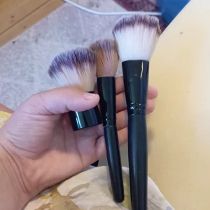 Set med tre sminkborstar - Säljer ett set med tre sminkborstar med svarta handtag och täta, mjuka syntetborst i beige och lila toner. En kabukiborste med kort handtag och två större borstar med långt handtag, perfekta för applicering av puder, bronzer eller rouge. Snyggt och stilrent set för din sminkrutin.