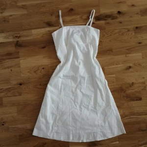 Vit sommarklänning med smala axelband - Vit klänning med smala axelband och rak halsringning. Klänningen är knälång med lätt A-linjeformad siluett och har ett tunt foder under. 