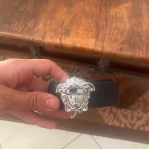 Svart Versace bälte med Medusa-spänne - Snyggt svart bälte från Versace med ett ikoniskt Medusa-huvud i silver som spänne. Bältet är brett och har en stilren design som verkligen sticker ut. Perfekt för dig som vill ha en statement-accessoar med lyxig känsla.