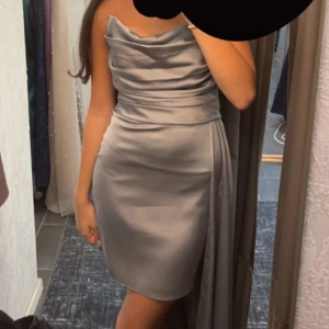 Silvergrå satin klänning med drapering - Snygg silvergrå kort klänning i satin med draperad överdel och axelbandslös design. Klänningen har en figurnära passform och ett elegant fall med extra tygdetalj på sidan. 