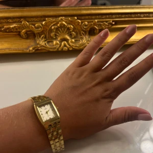 Guldfärgad klocka med länkarmband - Stilren guldfärgad klock med fyrkantig urtavla och klassiskt länkarmband. Urtavlan är vit med svarta visare och diskreta markeringar. Perfekt accessoar för dig som gillar en elegant och tidlös look.
