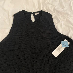 Svart stickad linnetopp från Stories  - Svart stickad linnetopp från Stories i storlek XL. Toppen har ett snyggt hålmönster, rund hals och knapp i nacken för en clean look. Ärmlös och perfekt för att styla med jeans eller kjol. Ribbad kant runt halsen och armarna ger extra edge.