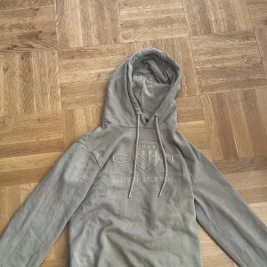 Beige hoodie från GANT med tryck - Säljer en beige hoodie från GANT i storlek XS. Tröjan har ett broderat GANT-tryck på bröstet, snörning i huvan och mjukt fleecefoder på insidan. Perfekt för chill dagar och har lång ärm och klassisk passform.
