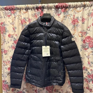 Svart Moncler pufferjacka herr - Snygg svart pufferjacka från Moncler med klassisk logga på ärmen. Jackan har quiltad design, dragkedja framtill och två fickor med dragkedja. Perfekt för dig som vill ha en stilren och varm jacka med modern look. Materialet är polyester och dun.