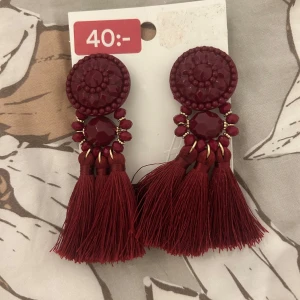 Mörkröda örhängen med tofsar från H&M - Snygga mörkröda örhängen från H&M med stora runda detaljer och glittriga stenar. De har flera tofsar som hänger ner och ger en cool och trendig look. Perfekta för att lyfta din outfit med lite extra färg och stil.