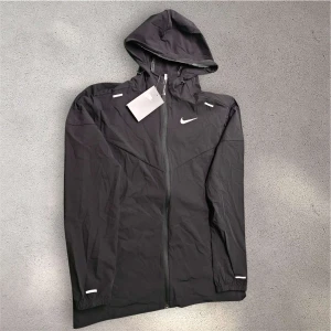 Svart nike Miler jacka - Snygg svart vindjacka från Nike med huva och hel dragkedja framtill. Jackan har vita detaljer och Nike-logga på bröstet. Lätt och smidig design, perfekt för aktiva dagar. Elastiska muddar och reflekterande inslag på ärmarna ger extra stilpoäng.