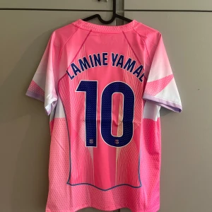 Barça Lamine Yamal rosa matchtröja - Snygg FC Barcelona fotbollströja med Lamine Yamal och nummer 10 på ryggen. Tröjan är rosa med vita och lila detaljer, har korta ärmar och coolt grafiskt mönster. Nike-logga, Barça-emblem och Spotify-logga på bröstet. Perfekt för dig som älskar fotboll och vill sticka ut.