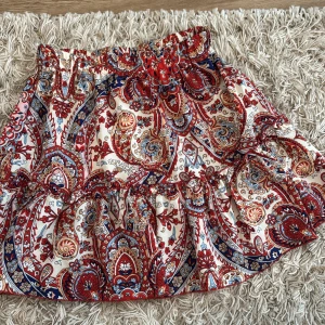 Mönstrad kortkjol från SHEIN - Säljer en färgstark kortkjol från SHEIN i storlek M. Kjolen har ett paisleyinspirerat mönster i rött, blått, beige och gult, med volangdetaljer och resår i midjan. Materialet är lätt polyester, perfekt för varma dagar och ger en bohemisk vibe.