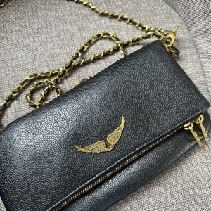 Svart axelväska Zadig & Voltaire - Snygg svart axelväska från Zadig & Voltaire i äkta skinn med guldfärgade detaljer och kedja. Väskan har ett coolt guldfärgat vingemblem på framsidan och dubbla dragkedjor. Perfekt storlek för mobilen och det viktigaste.