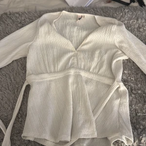 Vit omlottopp från Nelly - Säljer en vit omlottopp från Nelly i storlek S. Toppen har lång ärm, v-ringning och knytband i midjan. Materialet är ribbat och har en lätt struktur som ger en cool och modern vibe. Perfekt att styla med jeans eller kjol för en fräsch look.