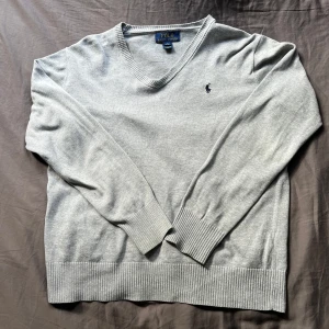 Grå v-ringad tröja från Polo Ralph Lauren - Snygg grå v-ringad tröja från Polo Ralph Lauren med klassisk broderad logga på bröstet. Tröjan har långa ärmar, ribbade muddar och nederkant. Perfekt för dig som gillar stilrena och tidlösa plagg. Den passar men på gränsen för mig som är 164 cm lång. Mvh Max