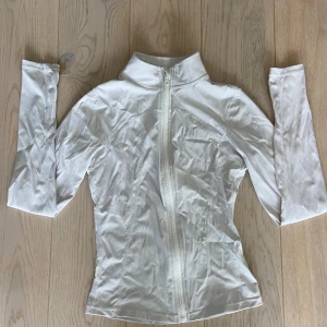 Vit sportig zip-jacka XS - Slimmad vit jacka med hel dragkedja och hög krage, tillverkad i stretchigt material (polyamid och elastan). Långärmad med tumhål i ärmsluten för extra komfort. Perfekt för träning eller chill dagar. Snygg och enkel design som sitter tajt mot kroppen.