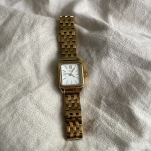 Guldfärgad fyrkantig klocka från Regal - Snygg guldfärgad klocka från Regal med fyrkantigt ur och vit urtavla. Länken är i metall med klassisk design och har ett smidigt spänne. Urtavlan har romerska siffror och stilrena detaljer. Perfekt för dig som gillar stilrena accessoarer.