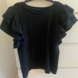 Svart ribbad topp med volangärmar - Snygg svart ribbad topp med breda volangärmar som ger en cool och trendig look. Tröjan har rund hals och är gjord i ett mjukt, stretchigt material som sitter skönt. Perfekt för dig som gillar något extra och vill sticka ut lite extra.