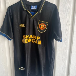 Manchester United Cantona 7 Umbro tröja - Retro Manchester United fotbollströja från Umbro med Cantona och nummer 7 på ryggen. Svart med gula detaljer och klubbmärke på bröstet. Kortärmad med blå mönstrade muddar och klassisk krage med gula ränder. Materialet är polyester, perfekt för match eller samling.