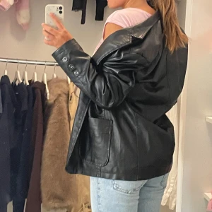 Svart oversized skinnkavaj - Säljer en svart oversized kavaj i skinn med klassisk krage och stora fickor framtill. Jackan har tre knappar på ärmsluten och en stilren, rak passform som ger en cool vibe. Perfekt för dig som gillar statement-plagg och vill sticka ut.